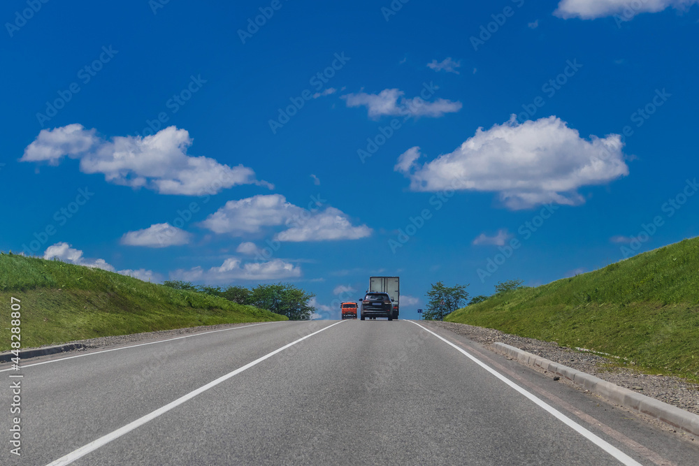 Summer rural landscape with green fields and highways with cars under a blue sky with white cumulus clouds in the countryside on a sunny day.