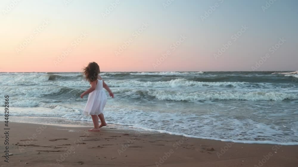 little girl child in a long sundress with long hair runs along the beach along the sea at sunset in summer. High quality 4k footage little girl child in a long sundress with long hair runs along the beach along the sea at sunset in summer. High quality 4k footage