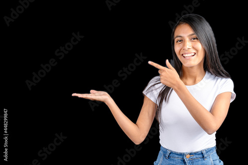 Bilde på lerret Beautiful Latin woman with white t-shirt on black background with her outstretched hand holding imaginary and the other hand pointing to the imaginary held item