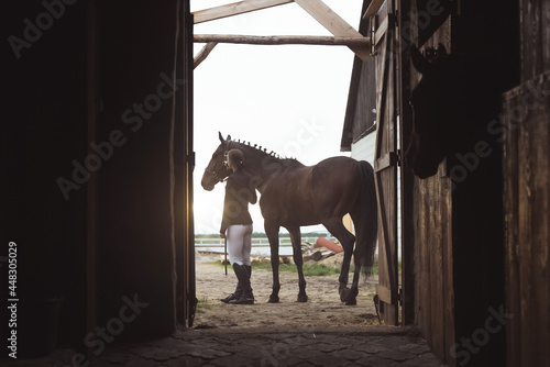 Horsewoman standing with her dark bay horse outside the stable. Posing for the camera. View from the door of the stable. Another horse can be seen in the stall. Daytime shot.