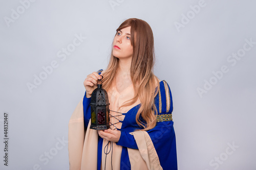 Portrait of a girl in a medieval blue dress with long hair and a lamp in her hands, posing isolated on a white background.