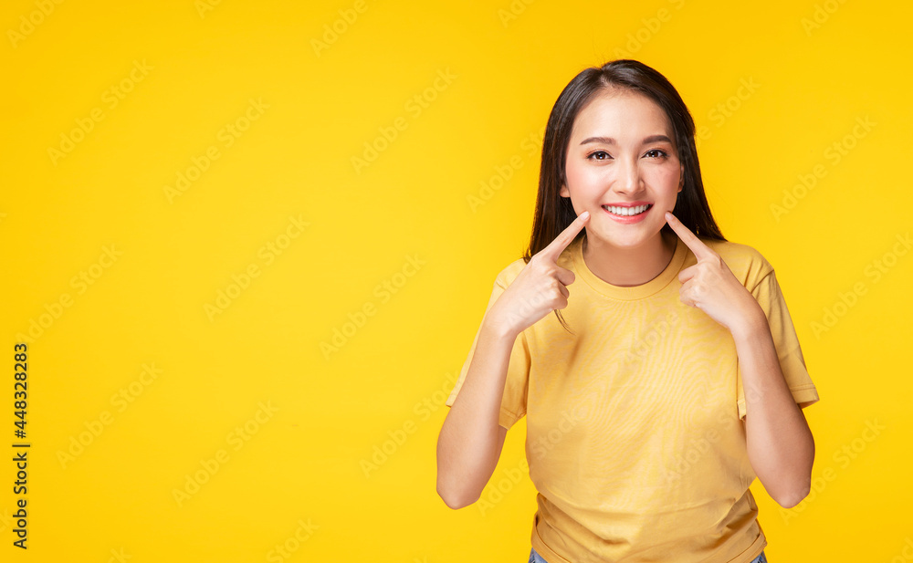 Cheerful young woman using finger point to her whitening teeth while standing over isolated yellow background. She showing her cleaning fresh toothy. Dental care and healthy concept. Cheerful young woman using finger point to her whitening teeth while standing over isolated yellow background. She showing her cleaning fresh toothy. Dental care and healthy concept.