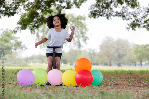 Childhood and relax concept - little African American curly hair girl useing a balloon inflator under big tree and dancing with enjoyment alone.