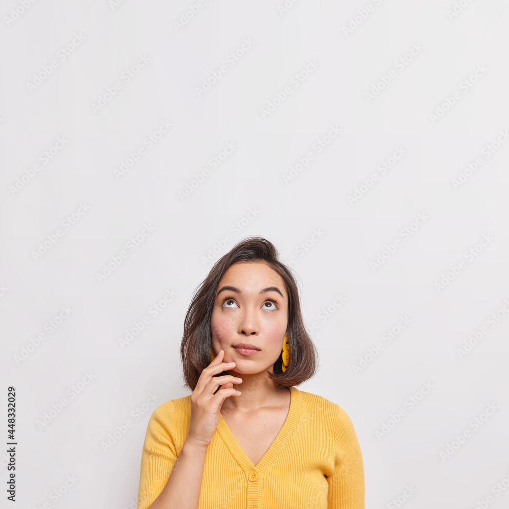 Vertical shot of dreamy thoughtful young Asian woman with dark hair focused above considers something wears casual yellow jumper isolated over white background copy space for your advertisement Vertical shot of dreamy thoughtful young Asian woman with dark hair focused above considers something wears casual yellow jumper isolated over white background copy space for your advertisement