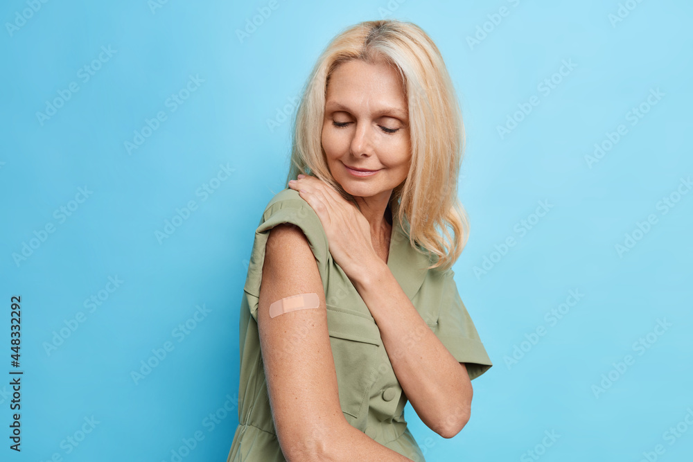 Horizontal shot of blonde European woman wears plaster on arm got vaccination to protects herself from coronavirus needs covid 19 immunization certificate poses against blue studio background Horizontal shot of blonde European woman wears plaster on arm got vaccination to protects herself from coronavirus needs covid 19 immunization certificate poses against blue studio background
