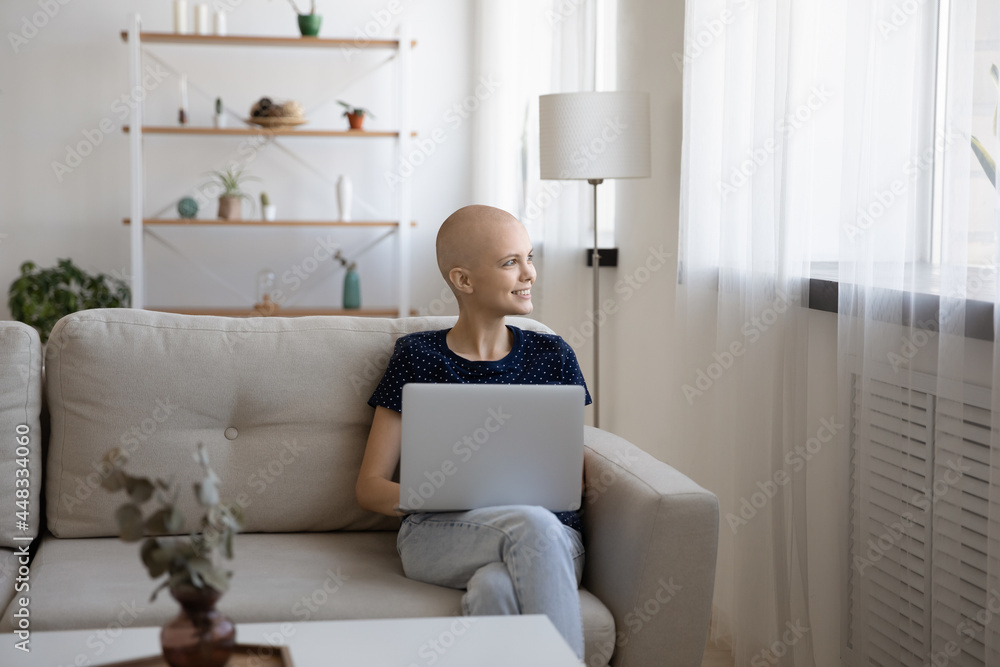 Dreamy young relaxed hairless cancer ill woman sitting on sofa with computer on laps, getting message with good news, looking in distance visualizing healthy future, enjoying carefree weekend time. Dreamy young relaxed hairless cancer ill woman sitting on sofa with computer on laps, getting message with good news, looking in distance visualizing healthy future, enjoying carefree weekend time.