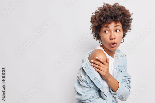Photo of shocked young African American woman keeps hand on sholder turns back gasps from wonder opens mouth from wonder wears denin shirt isolated over white background copy space for your text photo