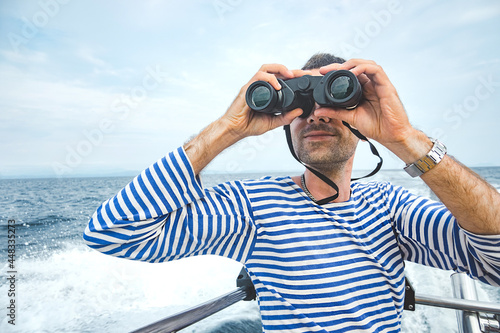 young man, sailor in striped jacket - vest with stubble sits on boat in sea and looks ahead through binoculars. sea navigation, summer tourism. travel content, selective focus