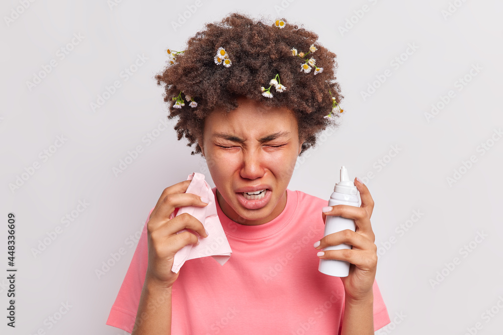 Sick unhappy curly haired Afro American woman uses nasal spray holds tissue feels unwell suffers from allergic rhinitis sneezes constantly isolated over white background. Remedy to cure allergy.