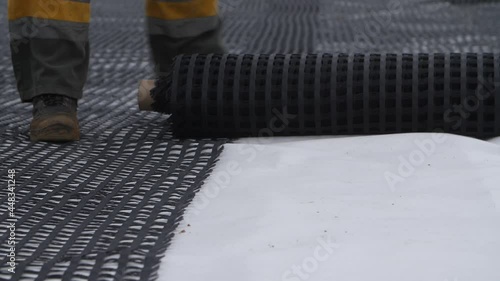 Close-up view of feet of construction worker or builder unrolling roll of plastic net, grid, geogrid or mesh for roadbed reinforcement over road surface. Geosynthetic material for highway base layer. photo