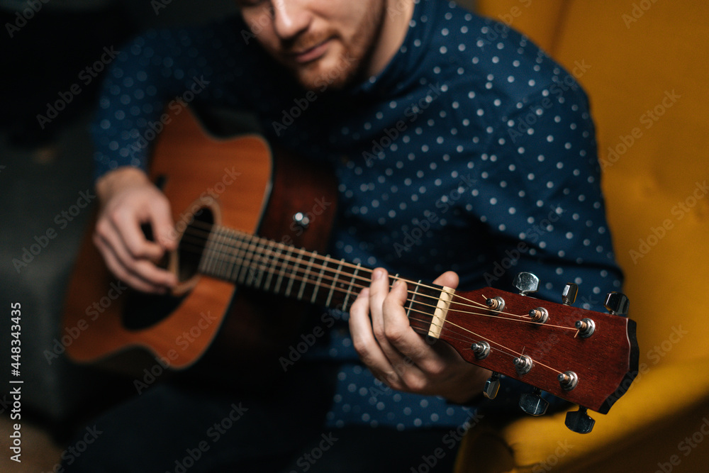 Obraz premium Close-up of guitarist singer male playing acoustic guitar sitting on armchair in dark living room, selective focus. Creative musician enjoying leisure activity in apartment.
