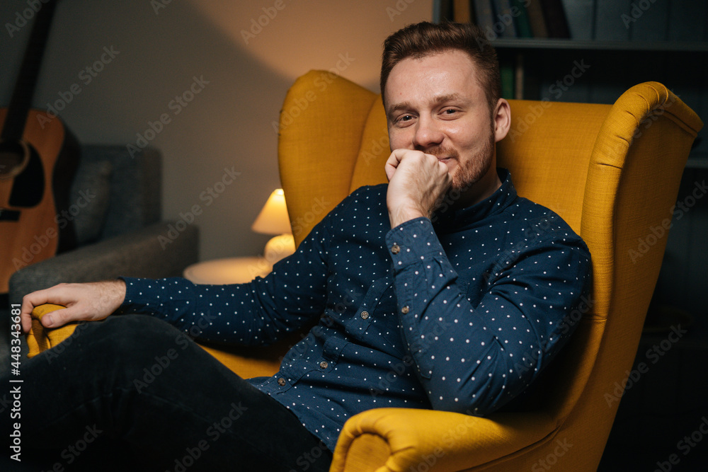 Portrait of smiling handsome man with beard in stylish shirt relaxing in luxurious yellow armchair, keeping hand in his chin, in dark cozy living room with modern interior, looking at camera. Portrait of smiling handsome man with beard in stylish shirt relaxing in luxurious yellow armchair, keeping hand in his chin, in dark cozy living room with modern interior, looking at camera.