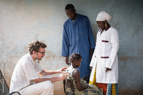 Two African physicians observing a young white doctor during auscultation of a small black girl's thorax during COVID screening photo