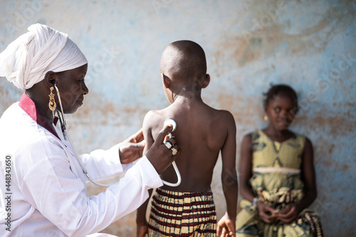 Smiling black doctor sitting in a classroom, auscultating one of two little African children with a stethoscope during COVID school screening