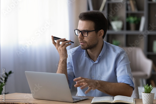 Modern tech in work. Focused male manager expert advisor using virtual assistant app on phone working by laptop in office. Young businessman hold phone by mouth use voice dialing or information search photo