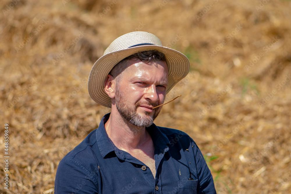 Portrait of a man 35-40 years old with a beard in a straw hat on a field with hay. Concept: a country boy, agriculture and summer seasonal work in the field, a straw in his mouth.