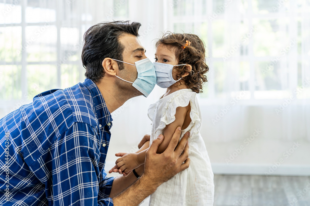 Caucasian father wearing face mask to avoid contagion  kissing daughter through medical mask, during covid-19 pandemic lockdown. Dad and baby daughter cuddling