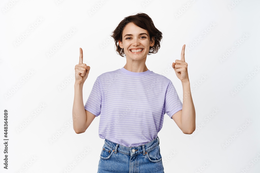 Cheerful smiling woman pointing fingers up, showing advertisement, empty space for logo, company service info, standing against white background Cheerful smiling woman pointing fingers up, showing advertisement, empty space for logo, company service info, standing against white background