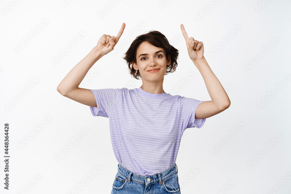 Smiling brunette woman pointing fingers up, looking pleased, showing advertisement, discount info, website link, standing against white background Smiling brunette woman pointing fingers up, looking pleased, showing advertisement, discount info, website link, standing against white background