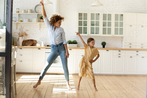 Happy mother and little daughter moving to favorite music in modern kitchen together, young mom teaching adorable kid girl to dance, family engaged in funny activity at home, enjoying leisure time photo