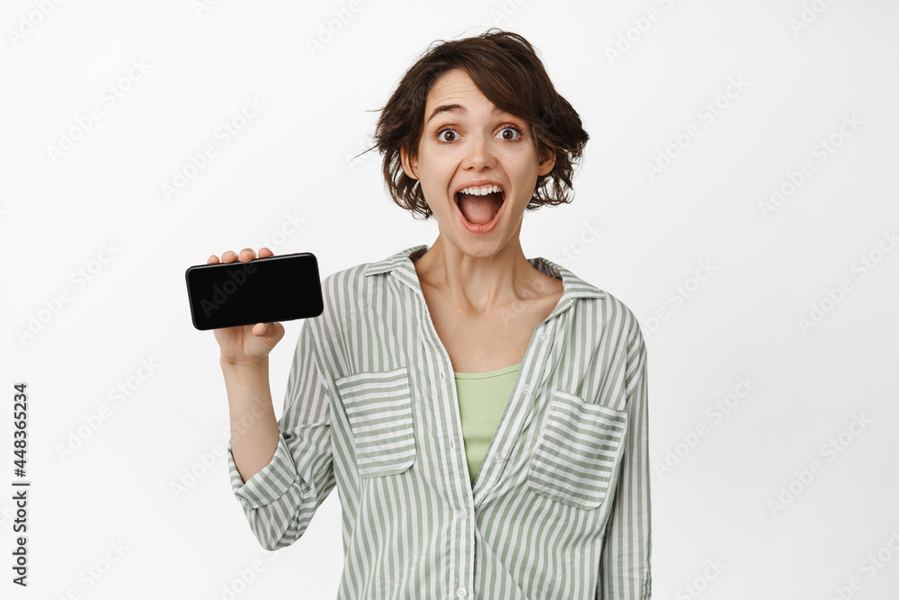 Portrait of surprised brunette woman showing horizontal smartphone screen, looking amazed, standing over white background Portrait of surprised brunette woman showing horizontal smartphone screen, looking amazed, standing over white background