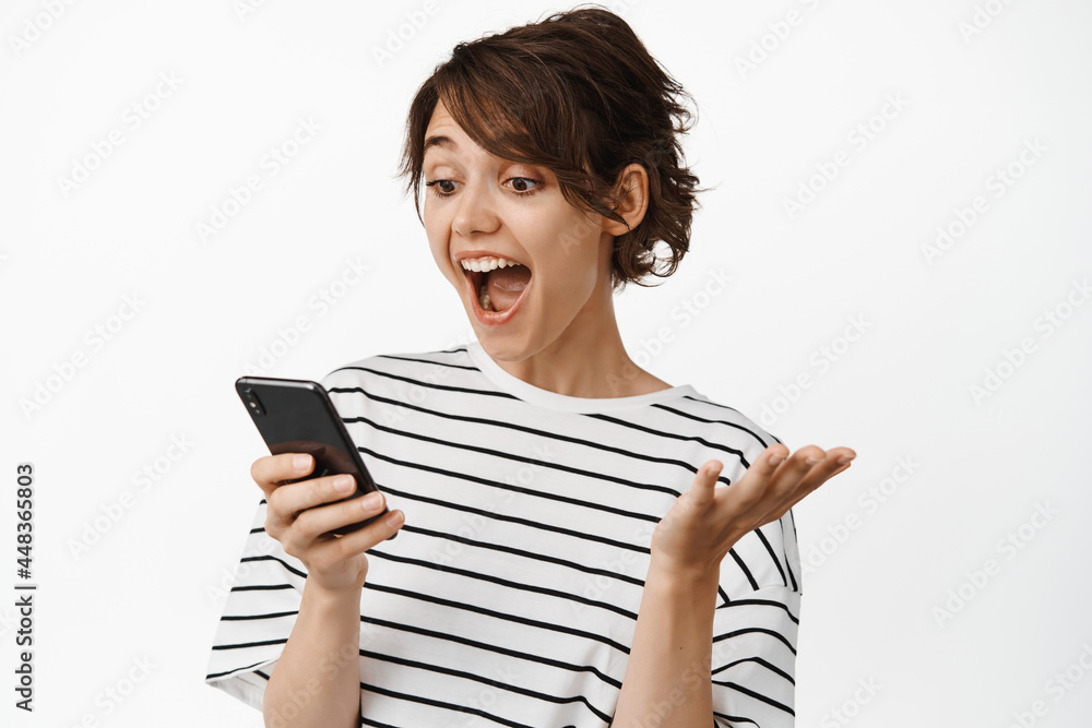 Naklejka premium Image of girl looking surprised at mobile phone screen, exciting with great news, notification on smartphone, standing over white background