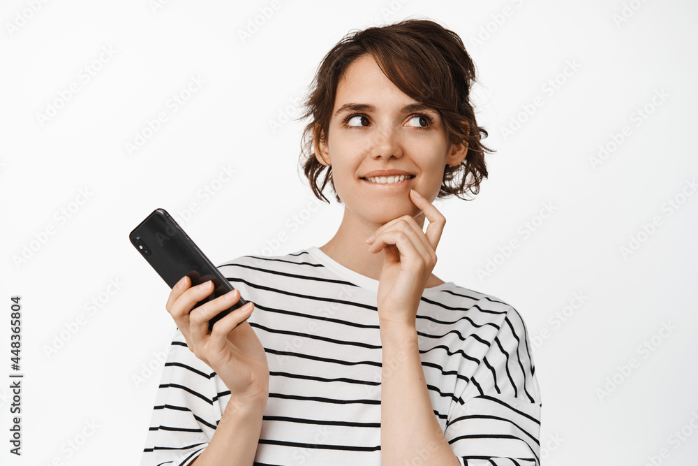 Portrait of young thoughtful woman holding mobile phone, smiling intrigued, tempted to try smth, having an idea, standing against white background thinking Portrait of young thoughtful woman holding mobile phone, smiling intrigued, tempted to try smth, having an idea, standing against white background thinking