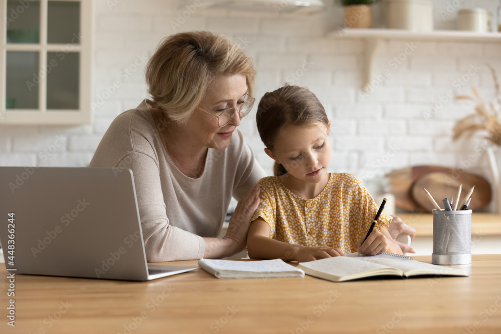 Caring mature grandmother in glasses helping to adorable granddaughter with school homework, sitting at table with laptop, studying, teacher babysitter teaching little girl, homeschooling concept Caring mature grandmother in glasses helping to adorable granddaughter with school homework, sitting at table with laptop, studying, teacher babysitter teaching little girl, homeschooling concept