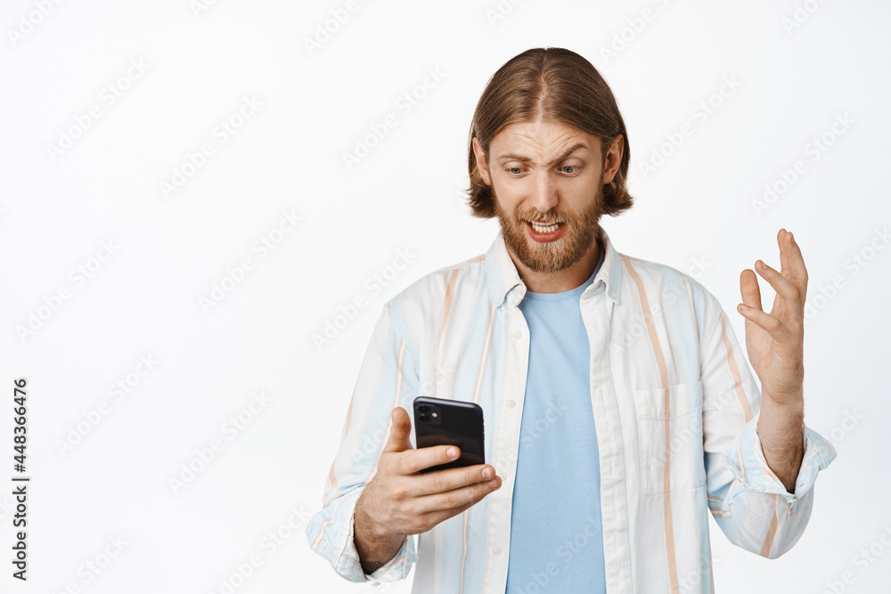 Frustrated and confused blond guy looking at mobile phone with worried face, reading bad news on smartphone, standing concerned against white background Frustrated and confused blond guy looking at mobile phone with worried face, reading bad news on smartphone, standing concerned against white background