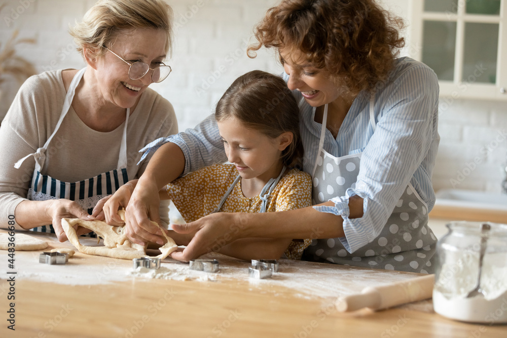 Happy three generations of women cooking homemade cookies using cutters, having fun with dough, cute little girl with mature grandmother in glasses and mother spending leisure time together Happy three generations of women cooking homemade cookies using cutters, having fun with dough, cute little girl with mature grandmother in glasses and mother spending leisure time together