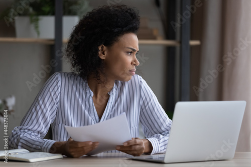 Unhappy young African American woman work online on laptop with paperwork feel unmotivated bored. Upset distressed biracial female consider document annoyed bothered at home office workplace.