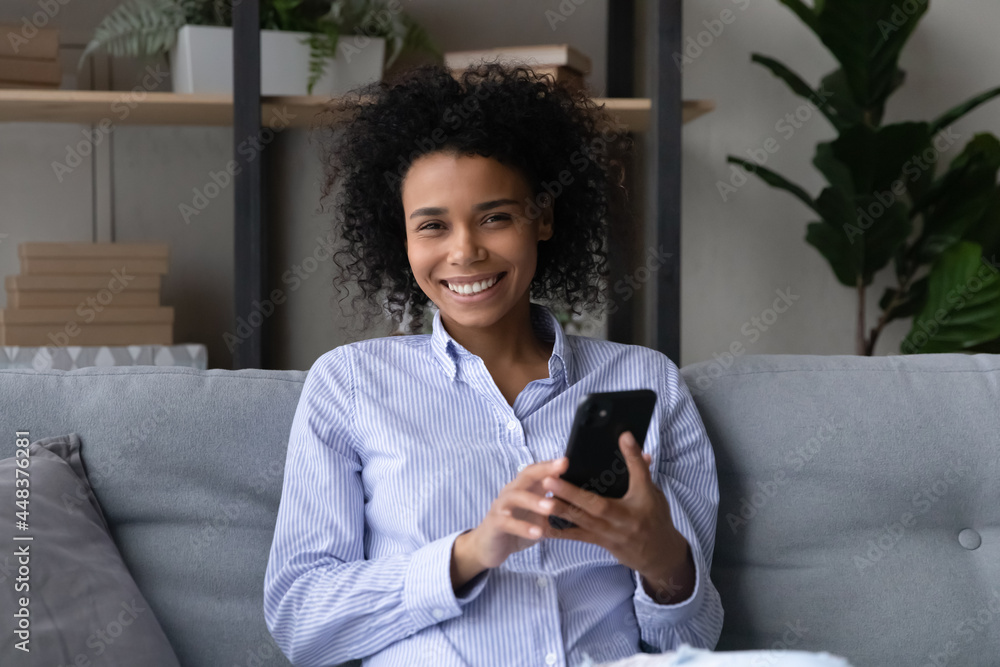 Portrait of smiling young African American woman relax on sofa at home using smartphone gadget. Happy biracial female talk speak on video call on cellphone, engaged in webcam virtual event on cell. Portrait of smiling young African American woman relax on sofa at home using smartphone gadget. Happy biracial female talk speak on video call on cellphone, engaged in webcam virtual event on cell.