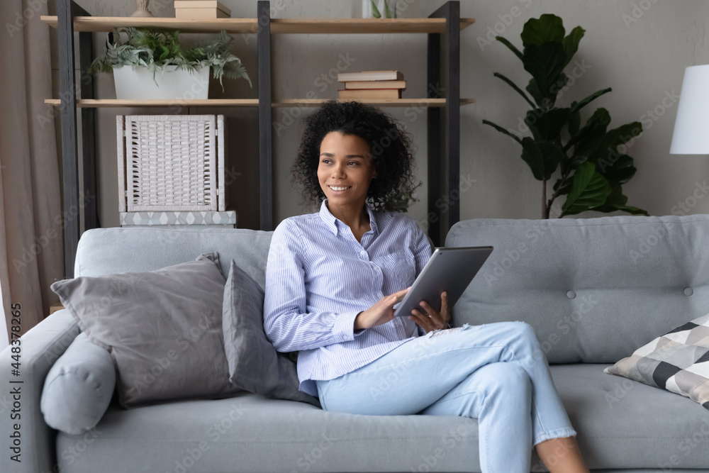 Smiling young African American woman sit relax on sofa in living room use modern tablet gadget look in distance. Happy millennial female rest at home browse internet on pad. Communication concept. Smiling young African American woman sit relax on sofa in living room use modern tablet gadget look in distance. Happy millennial female rest at home browse internet on pad. Communication concept.