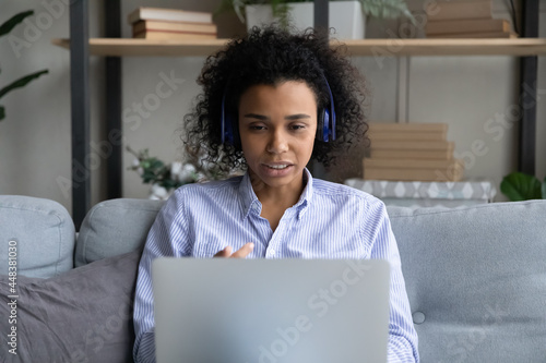 Young African American woman in headphones talk speak on video call on laptop with client customer. Biracial female have webcam online digital virtual event on computer. Communication concept.