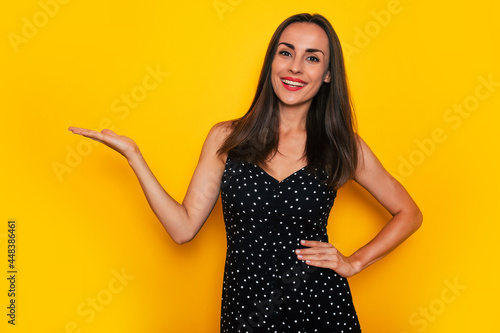 Happy beautiful young brunette woman in stylish dress shows empty palm isolated on yellow background © My Ocean studio