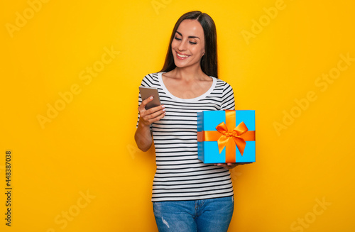 Attractive surprised excited young smiling woman is posing with big cool gift box in hands isolated on yellow background
