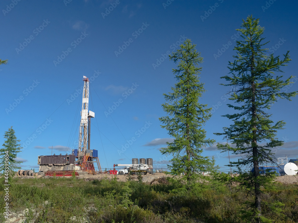 Landscape with a drilling rig in an oil and gas field. Infrastructure, communications and drilling equipment are visible. Around the rig there is summer northern taiga with green trees and blue sky