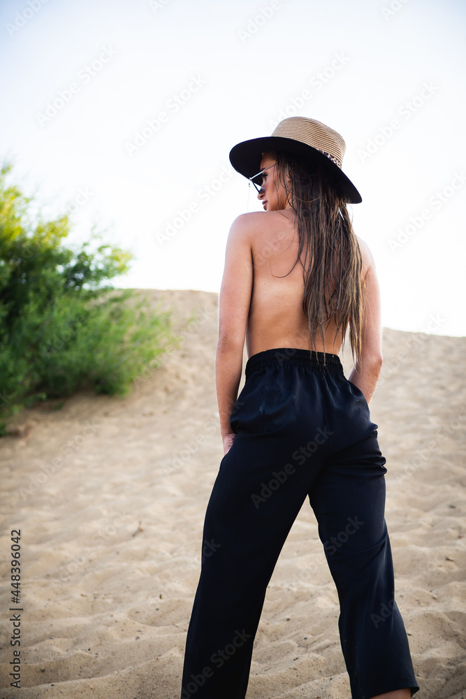 naked topless brunette model in black pants and a hat, posing on the sand in front of the camera.