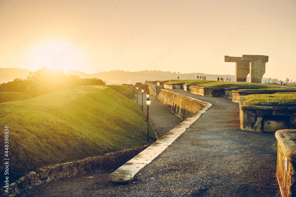 Promenade at sunny sunset of the Cerro de Santa Catalina next to the sea in the city of Gijón, in Spain. Between lampposts and green hills, you can see Elogio del Horizonte, by Chillida.