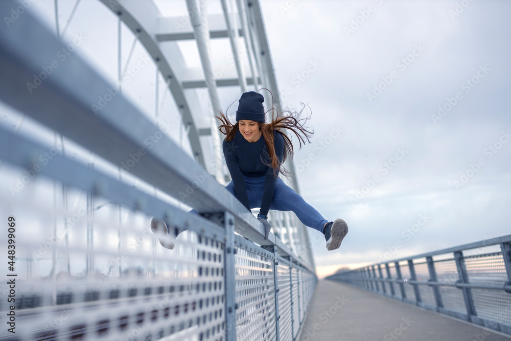 Young happy fitness woman in winter blue sportswear skips over bridge fence during cold winter day