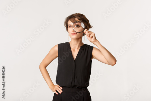 Intrigued stylish woman looking through magnifying glass, smiling thoughtful, standing over white background
