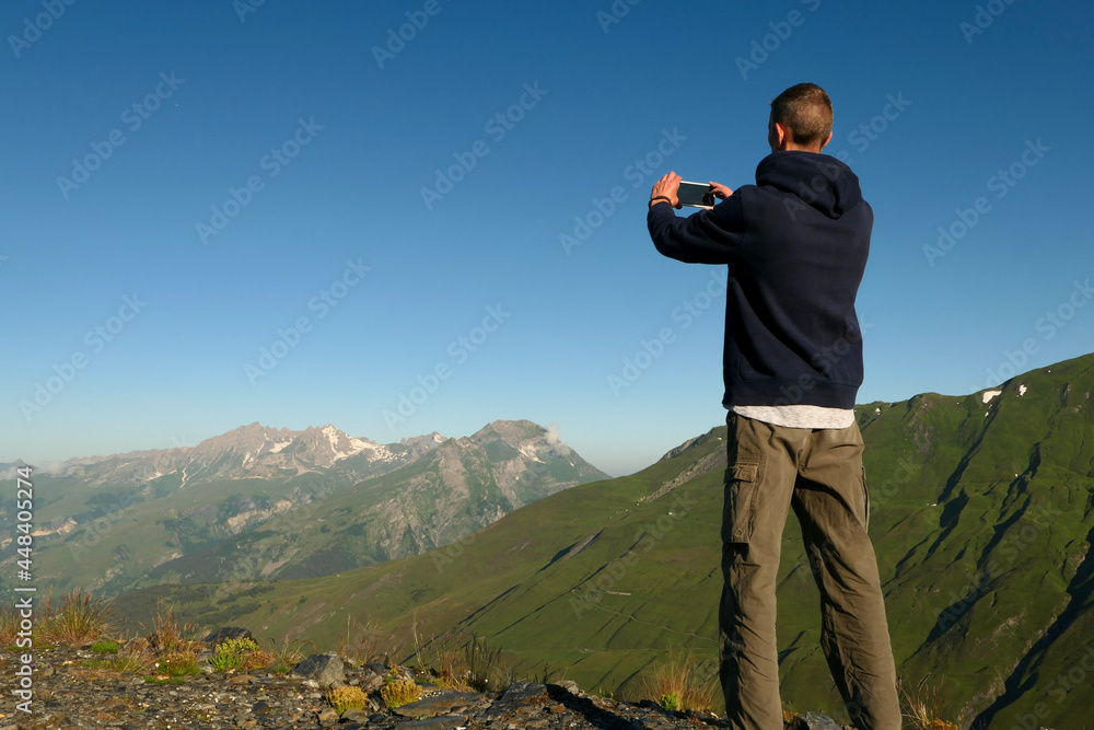 A man taking a picture with a smartphone in nature. Back hiker, at the top of a cliff, facing a mountain range
