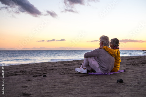 Portrait of couple of mature and old people enjoying summer at the beach looking to the sea smiling and having fun together with the sunset at the background. Two active seniors traveling outdoors..