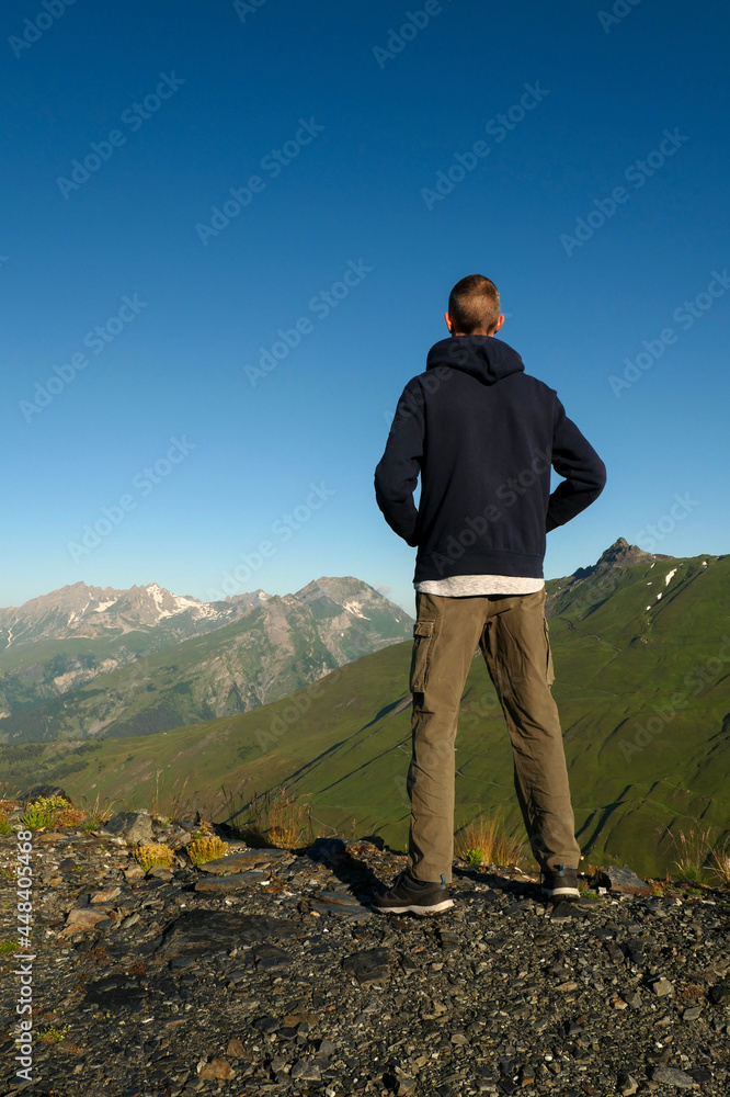 A man from the back, facing green mountains. A hiker contemplates the pastures and snow-capped peaks in summer at dawn.