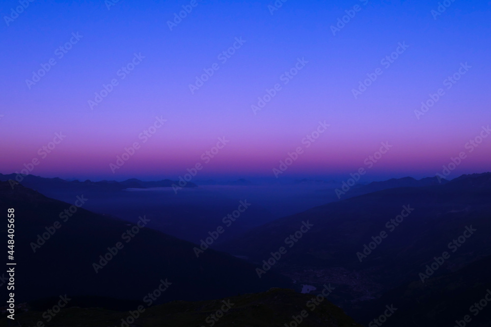 High-altitude view of a mountainous valley. Layer of clouds and pink mist above the mountains at dawn or sunset. High-altitude view of a mountainous valley. Layer of clouds and pink mist above the mountains at dawn or sunset.
