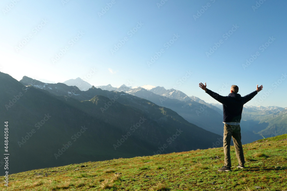 A man from behind, facing the mountain in summer. Hiker standing with arms outstretched up to the sky. Snowcapped mountain peaks in the background. Concept of success.