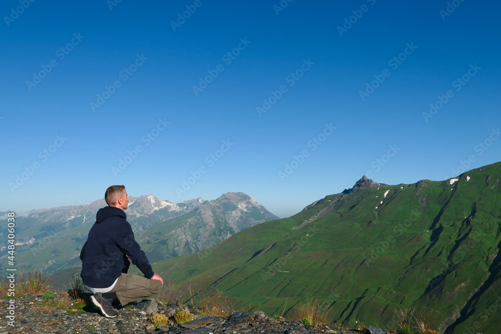 A man crouching on top of a mountain. Hiker facing a valley of the Alps in summer. A man crouching on top of a mountain. Hiker facing a valley of the Alps in summer.