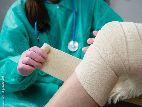 A nurse wearing medical gloves and a mask wraps an elastic bandage around the patient's leg and knee. Traumatologist applies elastic material to the patient in the physiotherapy room