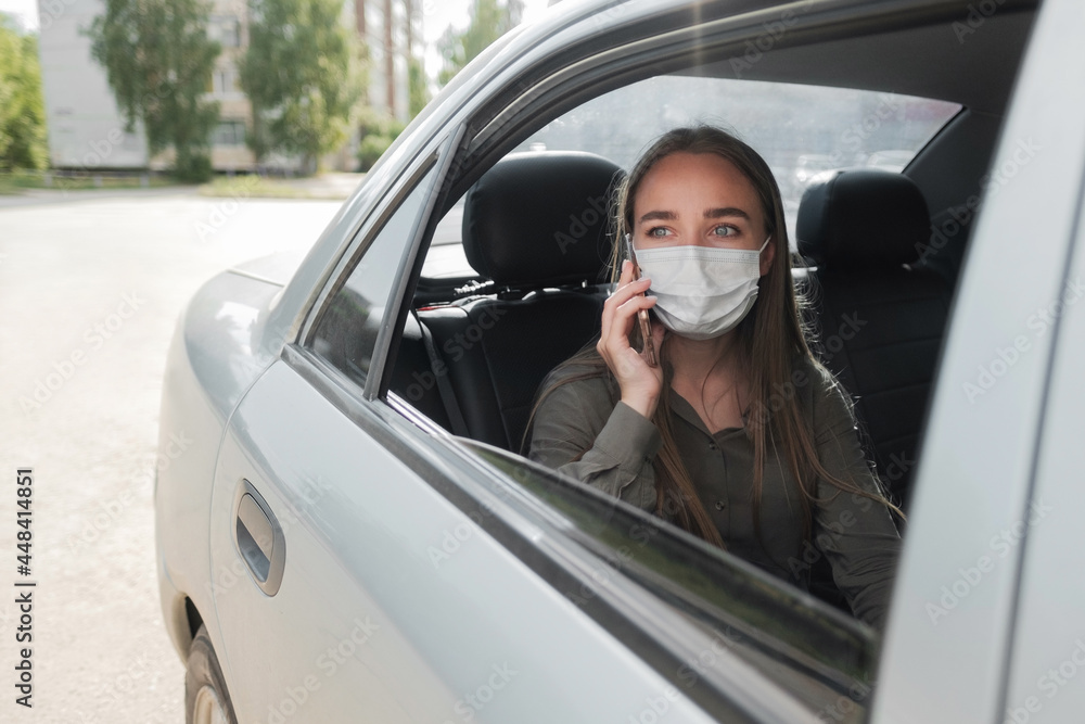 Naklejka premium A beautiful young girl in a mask is sitting in the car and talking on the phone. Protective mask against coronavirus.