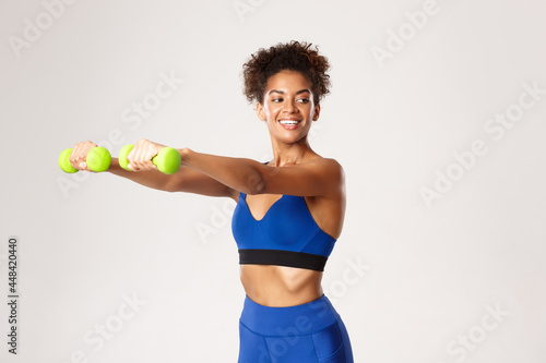 Concept of sport and workout. Healthy and happy african-american sportswoman in blue gym outfit, exercising with dumbbells against white background
